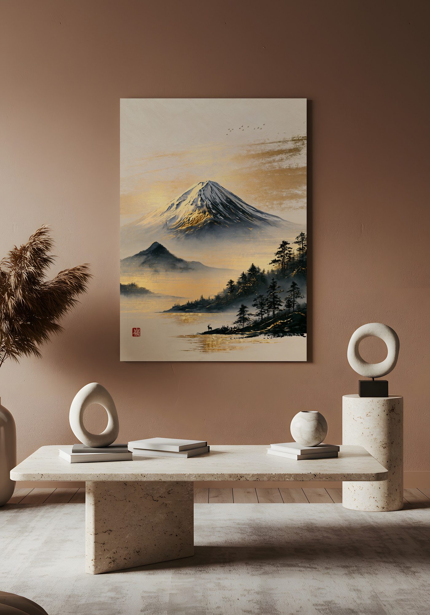 Mount Fuji, Brushed in Gold: a snow-capped volcano under a sky streaked with gold, above a lake that catches the gold. Dark pines stand on the shore at the right, and a small stag stands at the water's edge below them. A red seal sits in the lower left. The print hangs on a warm clay-colored wall above a low stone table. White ring sculptures, a round vase and small stacks of books stand on the table and on a stone pedestal at the right, and dried pampas grass rises on the left.